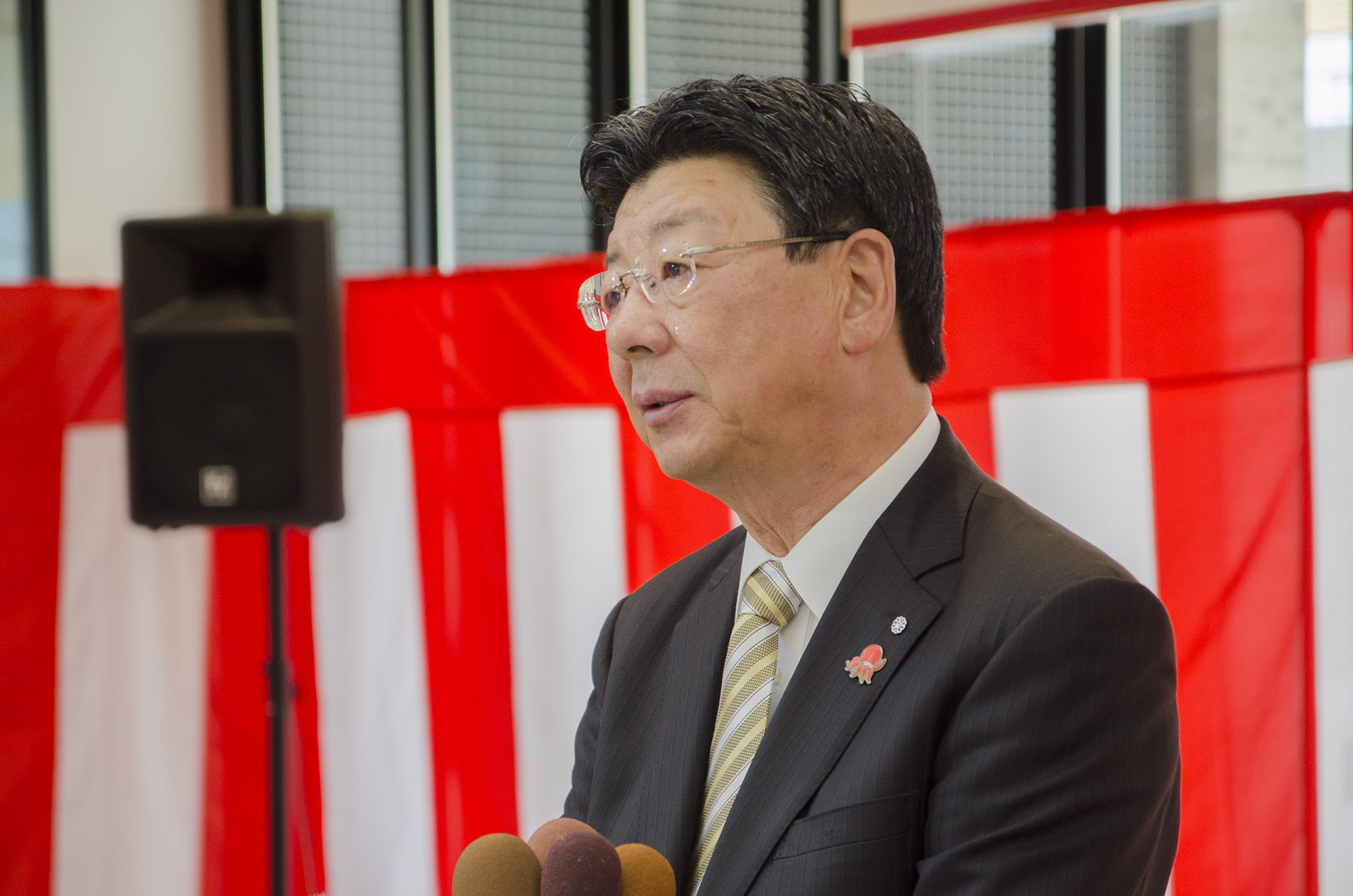 紅白の幕が張られた式典会場で、スーツ姿の佐藤仁町長がマイクの前で涙を浮かべながら話をしている写真
