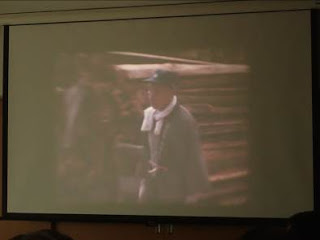 積み上げられた木材の前でタオルを首に巻き、法被を着た男性が作業をしている様子がスクリーンに映し出されている写真