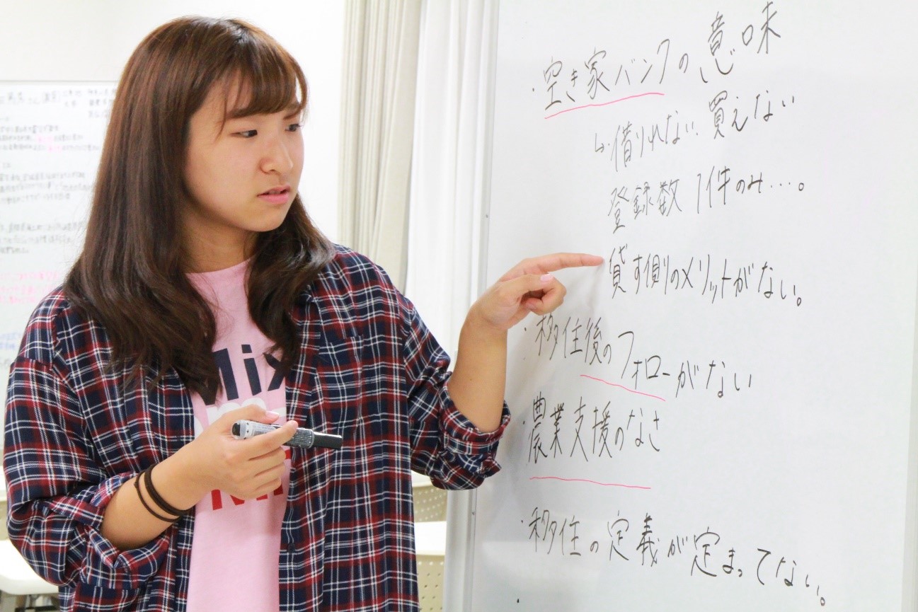 女性がホワイトボードの前に立ち、左手でボードの一部を指さしながら何かを説明している様子が写っている写真