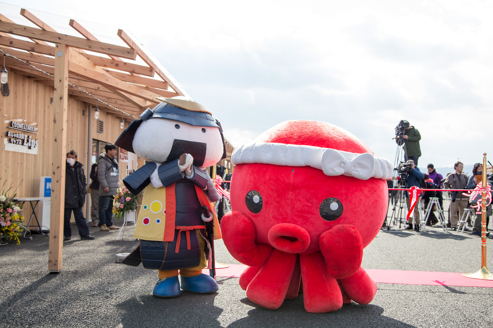 報道陣や見物客が見える商店街の入口付近で、赤いオクトパス君の着ぐるみと、顔がおむすびの形をした鎧を着た武士風のむすび丸の着ぐるみが並んでポーズをとっているマスコットキャラクターの写真
