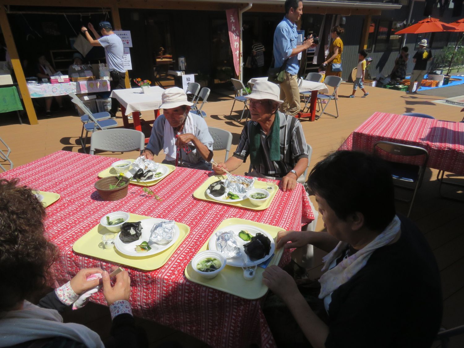 赤いテーブルクロスがかけられた屋外のテーブルで数人の高齢者が食事をしている様子を写した写真