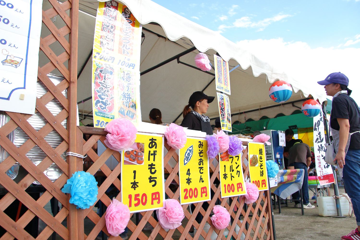 「あげもち150円」「みそおでん200円」などと書かれた手書きのポップが貼られた屋台の前で、販売する2人の女性スタッフが話をしている写真