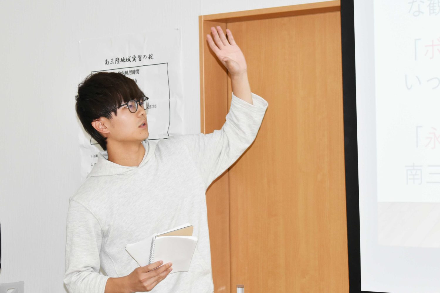 男性が室内でノートを右手に持ち、もう一方の手を挙げながらプレゼンテーションをしている様子が写っている写真