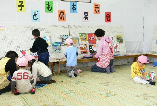 白地の壁に貼られた「子どもの本展示会」の文字の下で、複数の幼児と大人たちが床や低い台の上で絵本を選んだり読んだりしている様子の写真