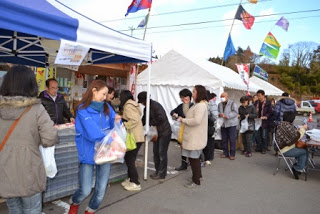 テントの前に並んだ来場者たちが、笑顔で会話をしながら地元の名物を買い求めている様子の写真