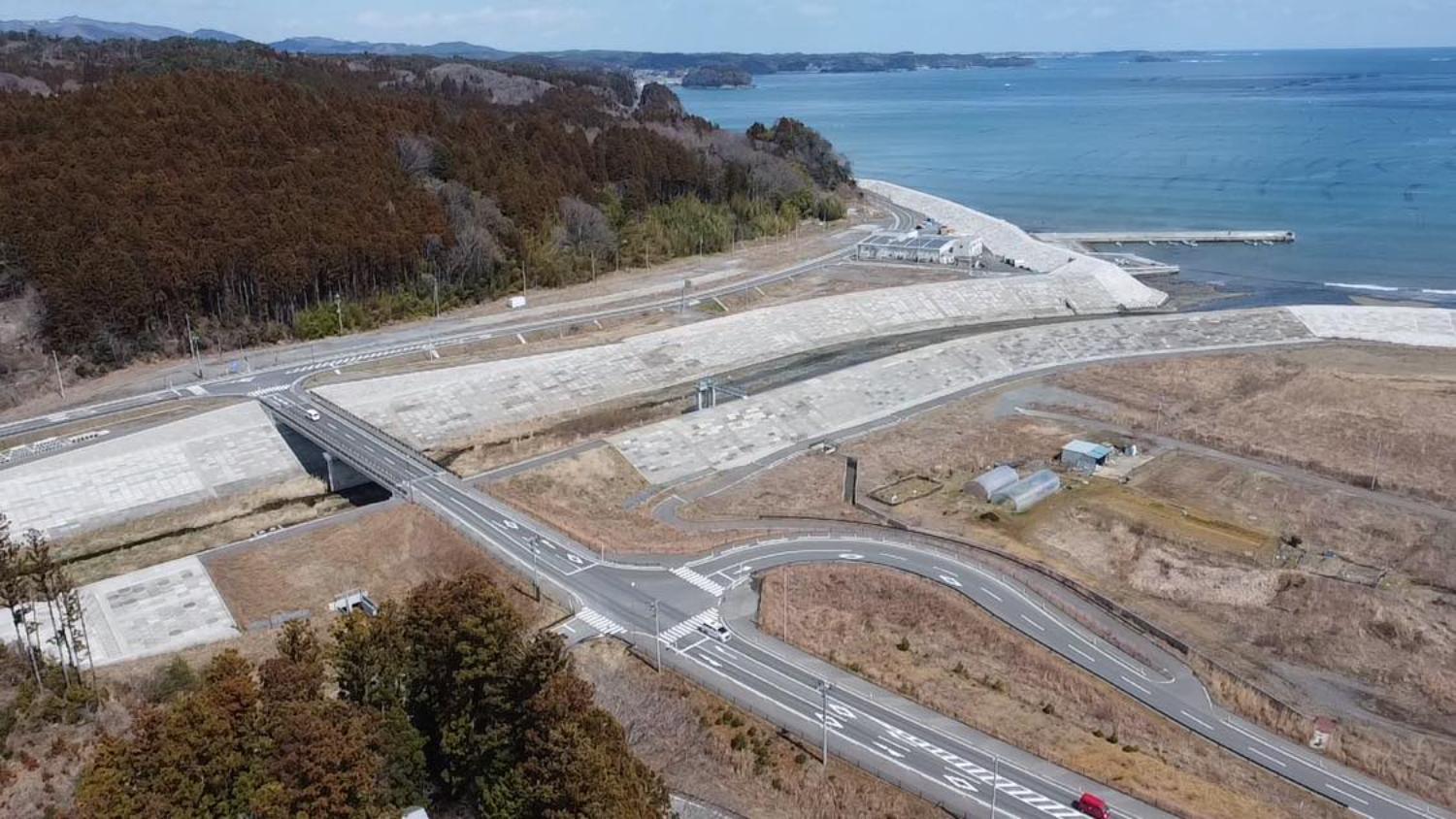 右手に広がる青い海と突き出した防波堤、手前には新しく整備された堤防と、それに沿って曲がりくねった道路や小さなビニールハウスが見え、海岸と内陸の境界がはっきり分かるような構図になっている海沿いの復興地域の昼間の風景を高台から定点カメラで撮影した写真