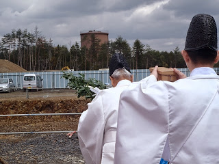 白装束を着た神職の男性が2人一列に並び、前の男性が手に榊を持ち、後ろの男性が木の箱を持ち、建設予定地で神事を行っている写真