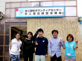 看板が設置され簾が立てかけられた南三陸町ボランティアセンターの入口前で、5人のスタッフがピースサインをして並んでいる写真