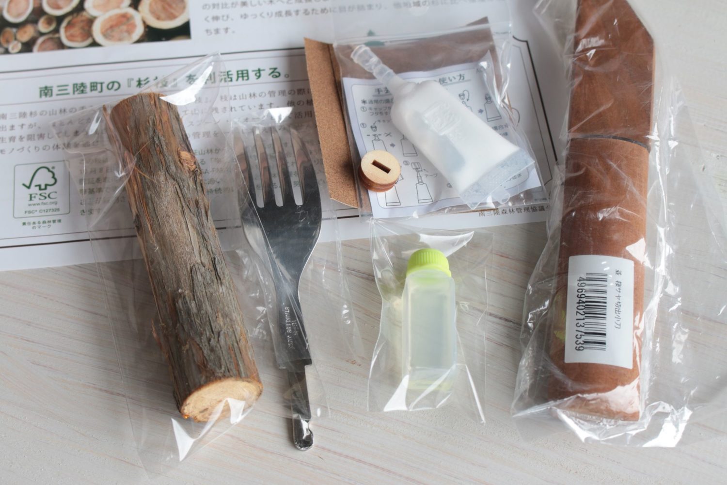 杉の木の枝、フォークの金具、接着剤やオイルなどの道具一式が袋に入れられ、木のカトラリーを手作りするためのキットが並べられている写真