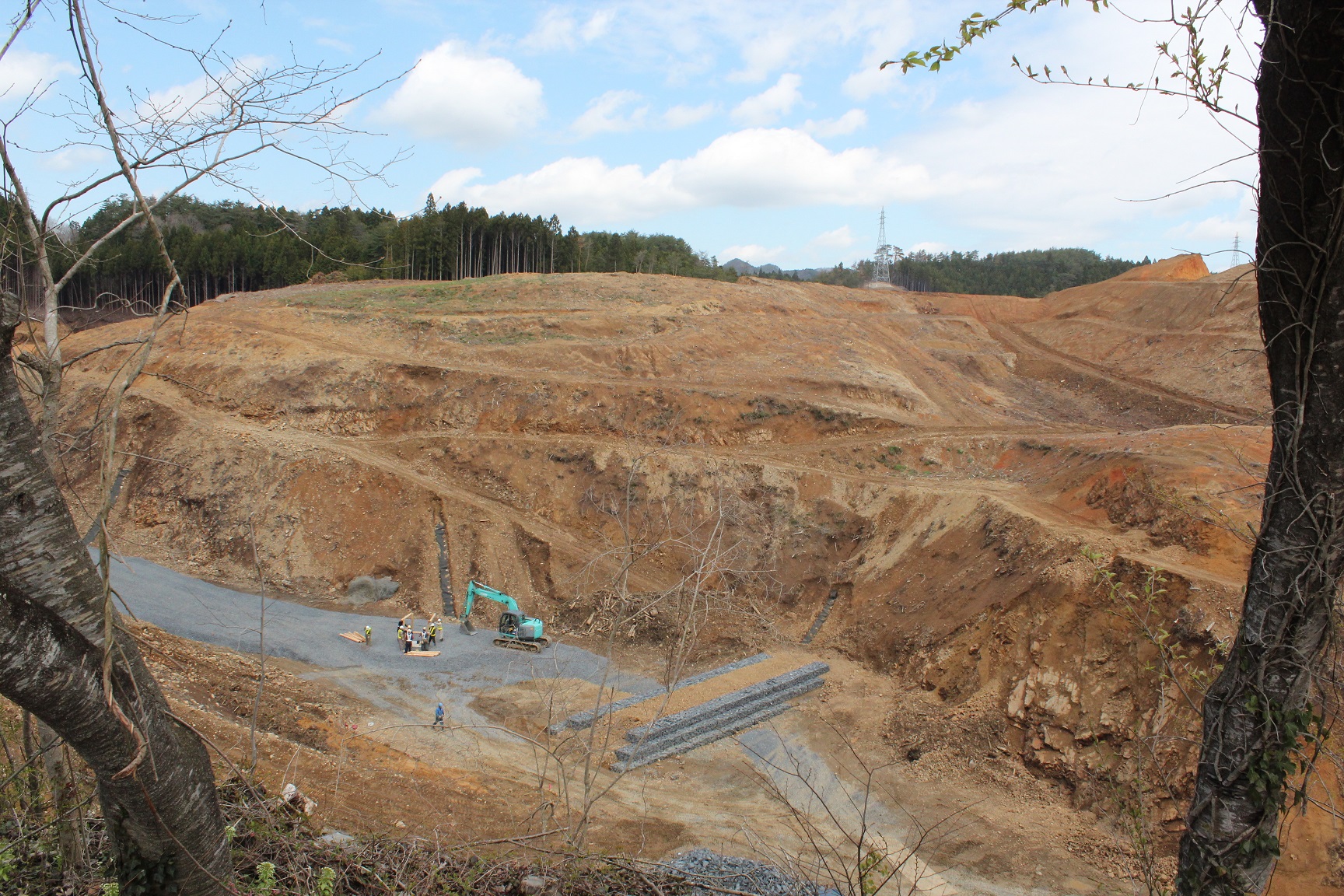 整地が進む広い造成地の麓に1台のショベルカーが停まっており、複数の作業員が木材を使って作業をしている様子の写真