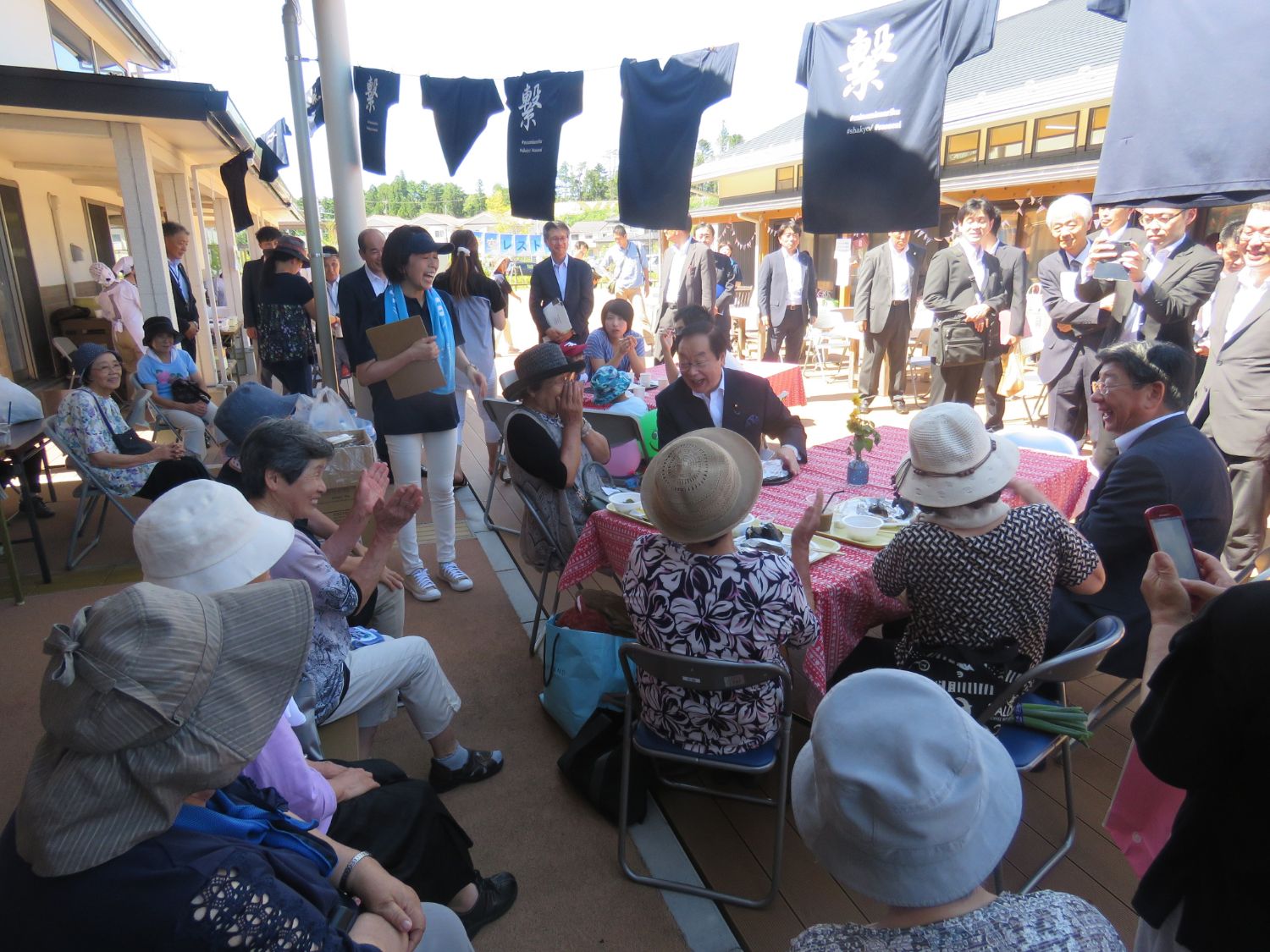 屋外のウッドデッキに設けられた赤いテーブルクロスのテーブルを囲んで多くの高齢者や来賓が笑顔で語り合い、上空には「繋」などと書かれた紺色のTシャツが飾られ、周囲には見守る人々や写真を撮る人の姿も見られる、和やかで交流に満ちた様子の写真