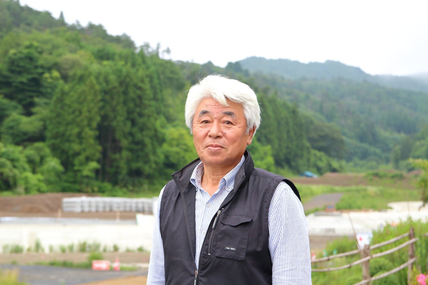 緑豊かな山あいの農村地帯で、白髪の男性がカメラに向かって微笑んでいる様子をアップで撮影した写真