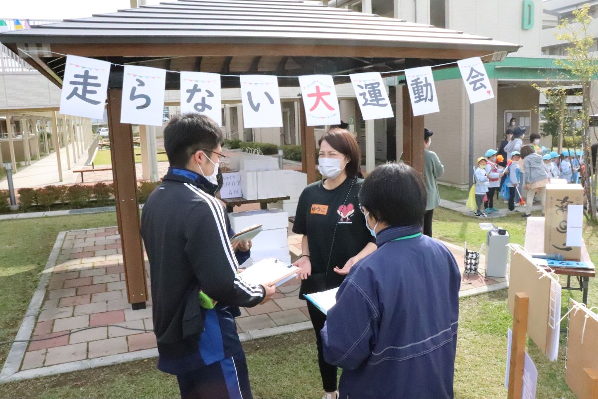 「走らない大運動会」と書かれた旗が掲げられた東屋の前で、マスクをつけたスタッフ三人が手に資料を持って話し合いをしている様子の写真