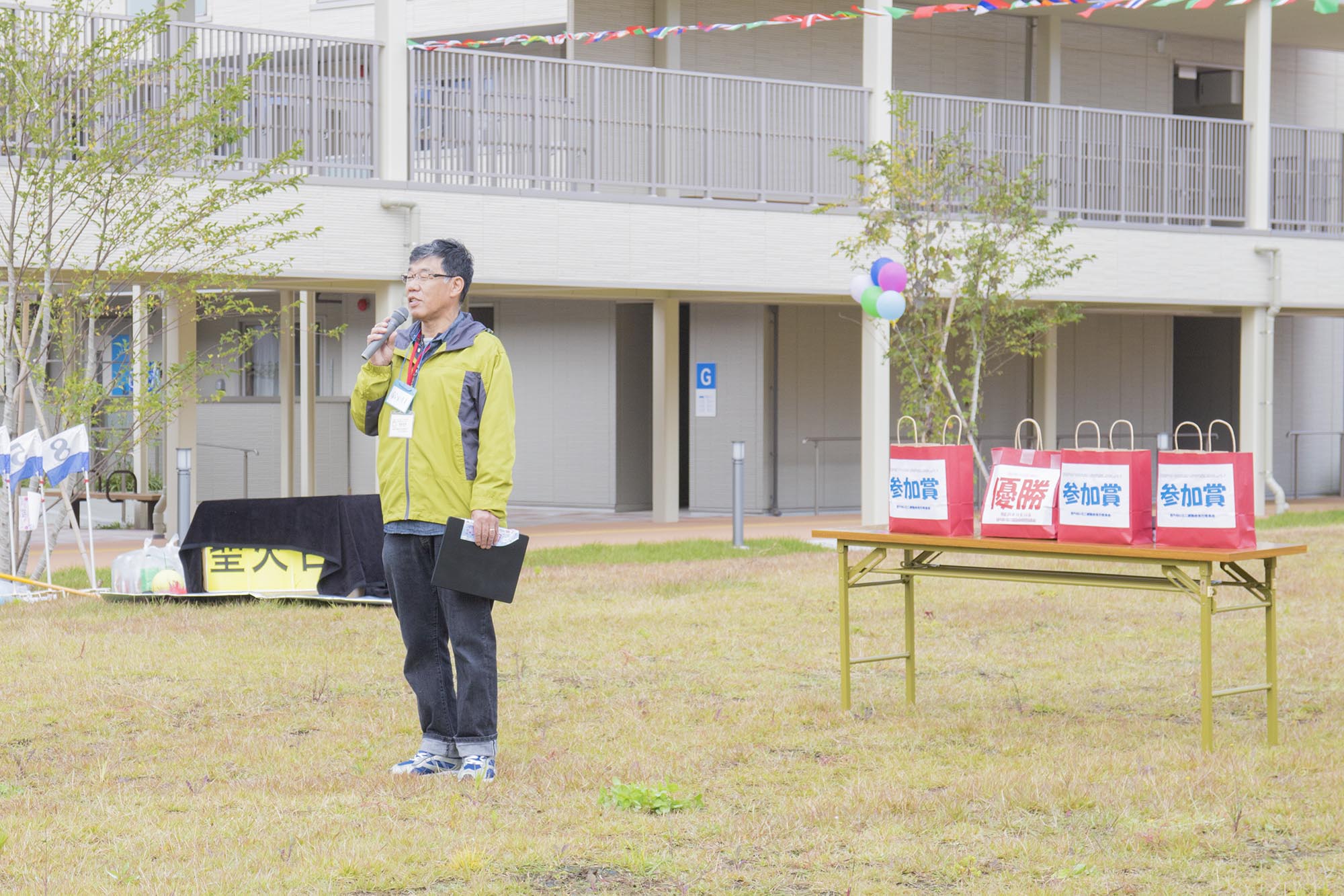 男性が屋外の広場でマイクを持って話しており、近くのテーブルには「参加賞」や「優勝」と書かれた赤い紙袋の景品が置かれた写真