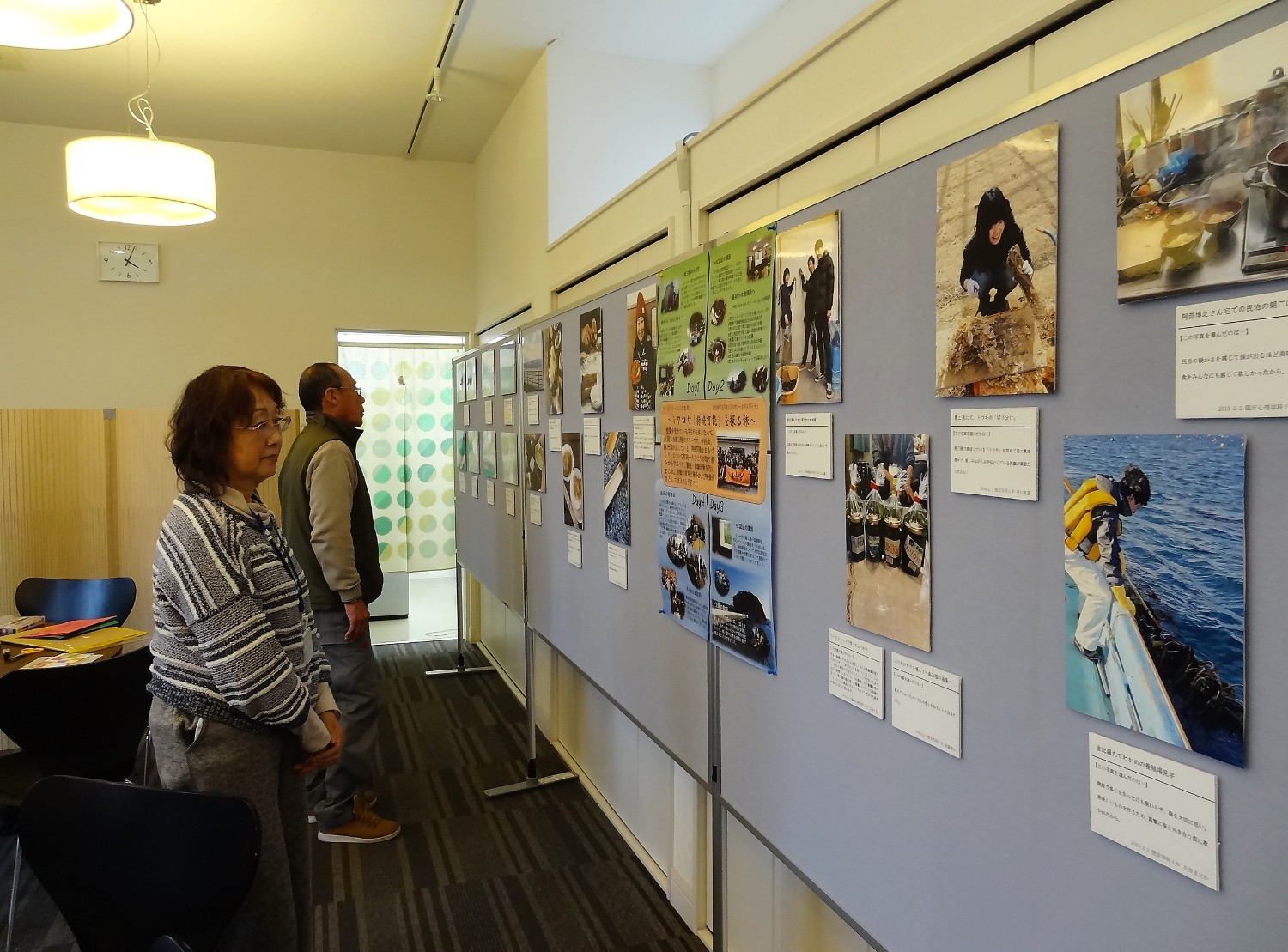 展示会の会場で、2名の方が壁に貼られた複数の写真や説明文をじっくりと見ている様子を写した写真