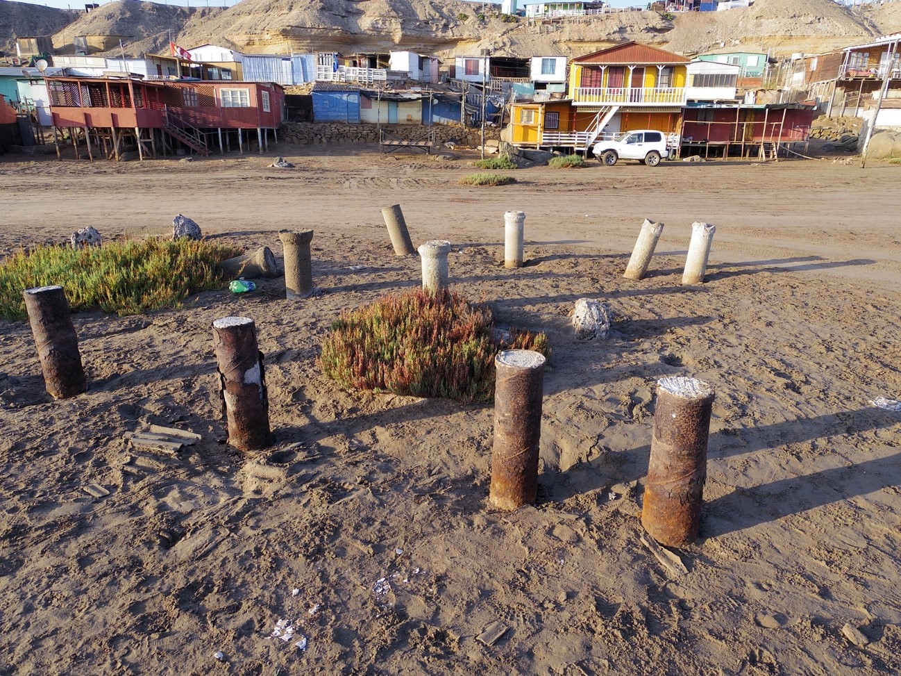 砂地に複数の古い柱の断片が立っており、その奥にカラフルな家々が並んでいる写真