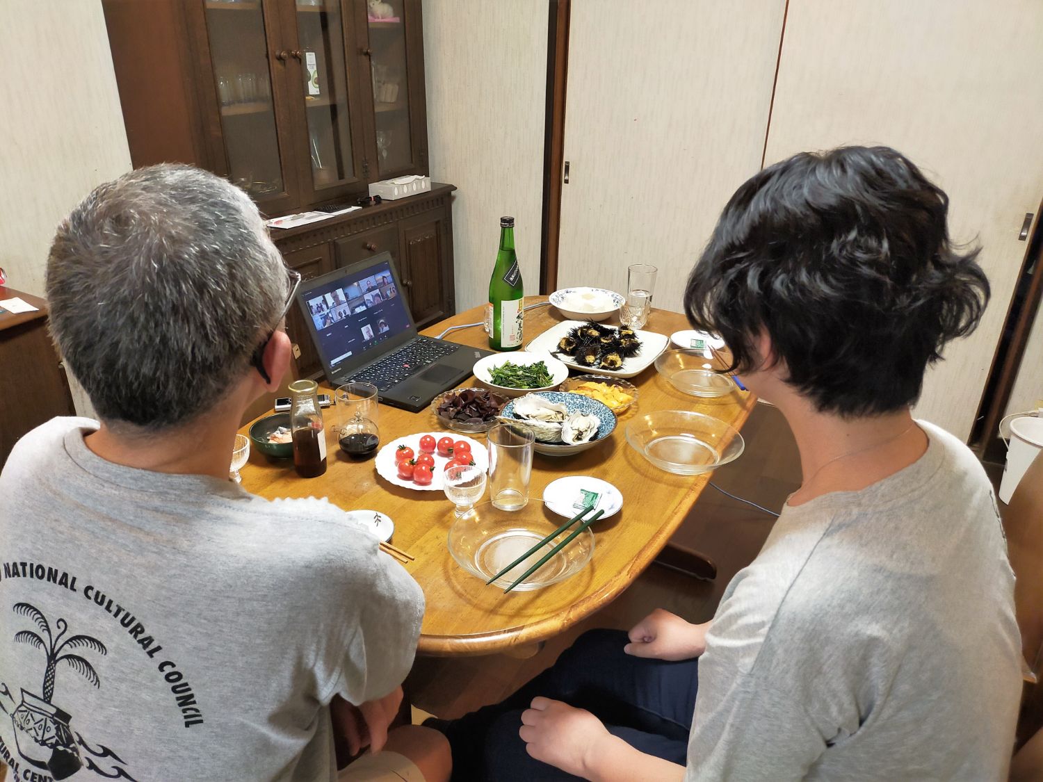 グレーのTシャツを着た男女が背中を向けて並んで座り、パソコンの画面を見ながら日本酒と海産物中心の料理を囲んでいる写真