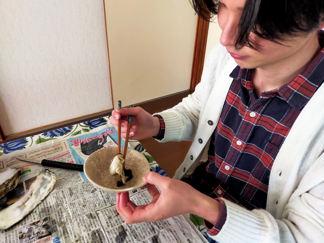 新聞紙が敷かれたテーブルの上で、チェックのシャツと白いカーディガンを着た男性が、開けた牡蠣の身を皿に移している様子の写真
