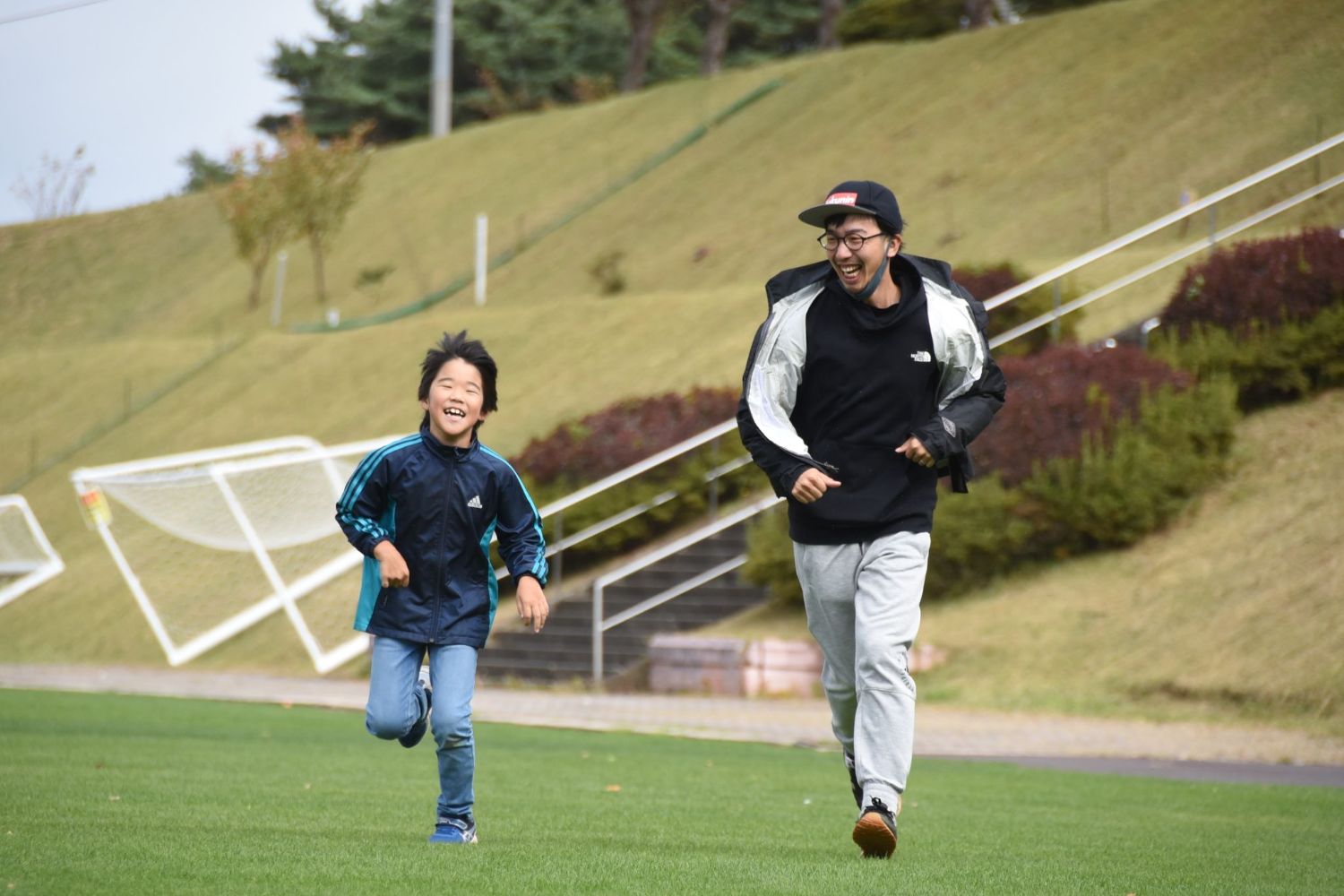 小学生くらいの男の子と大人の男性が一緒に横に並び芝生の上を笑いながら走っている写真
