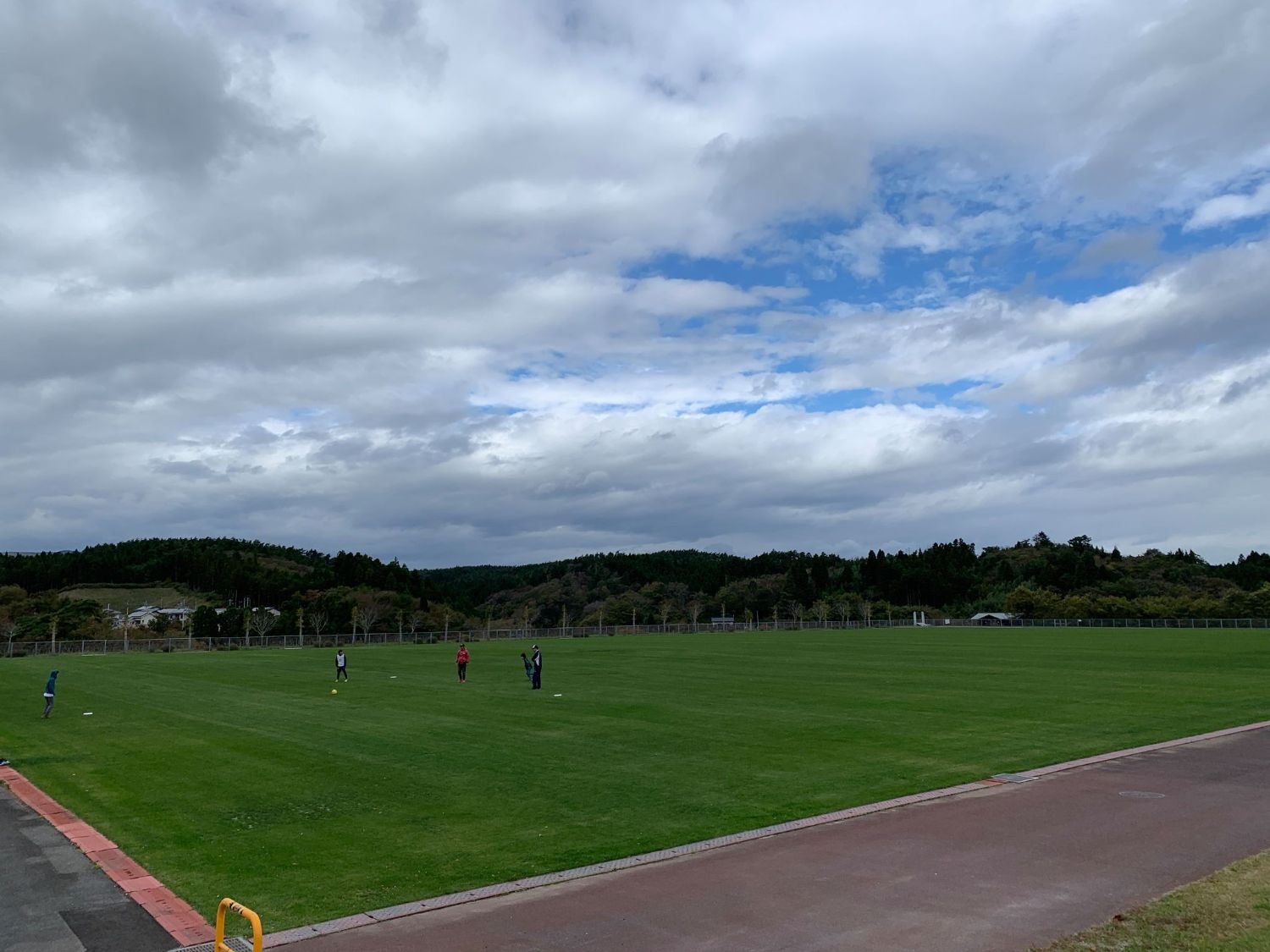 芝生の広場の中央で数人がフリスビーをしており、周囲には山や林が見え、空には雲が広がる写真
