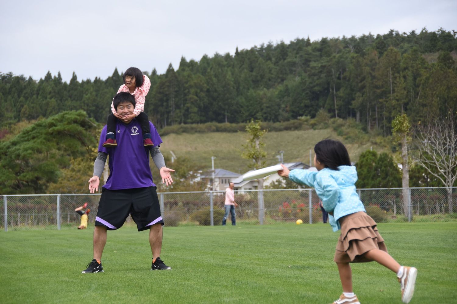 男性が女の子を肩車して立ち、向かい側で別の女の子がフリスビーを元気よく投げようとしている写真