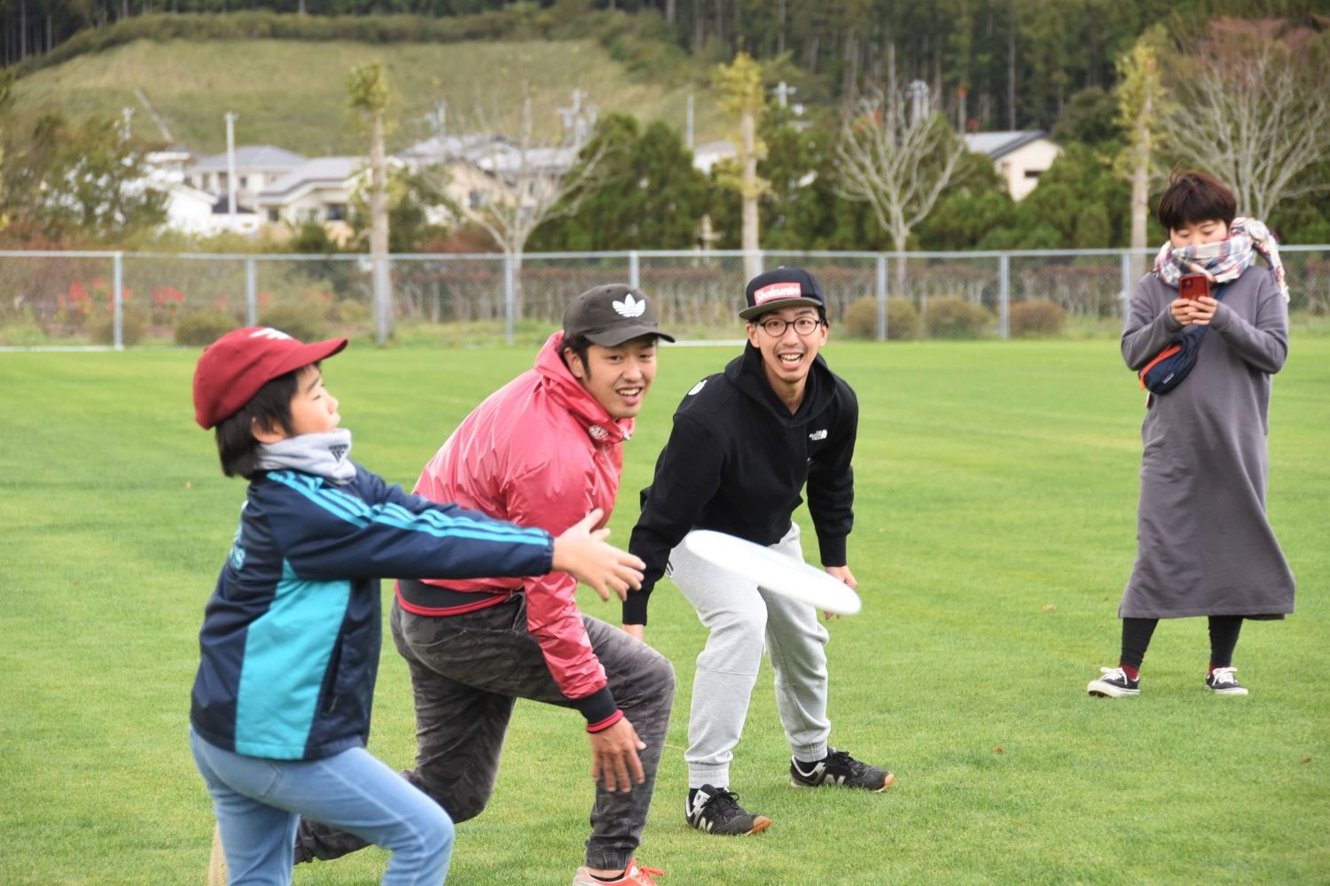 フリスビーを受け止めようとしている男の子と、周囲で一緒に動きながら笑っている二人の男性、そしてスマートフォンで撮影している女性が写っている写真