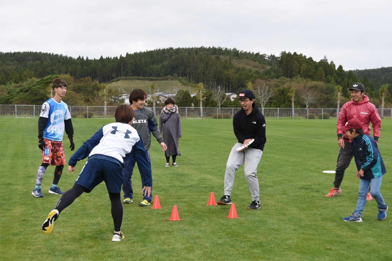 芝生の上を赤いコーンを囲んで投げられたフリスビーを男性が受け止め、周りの方の笑顔で見ている写真