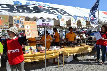 黄色い服を着たスタッフが立つ屋台では、「味わい開発室」と書かれた看板の下で様々な料理が販売されている、にぎやかな雰囲気の写真