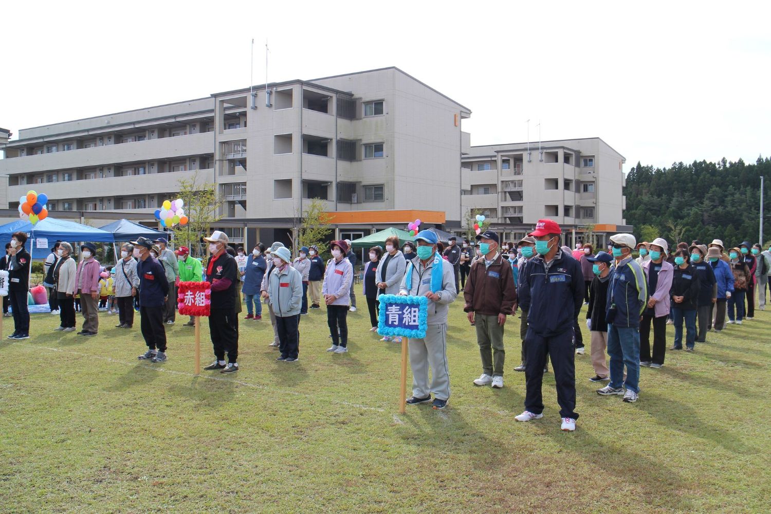 多くの大人がマスクをし先頭に立った赤組、青組のプラカードに分かれ整列している写真