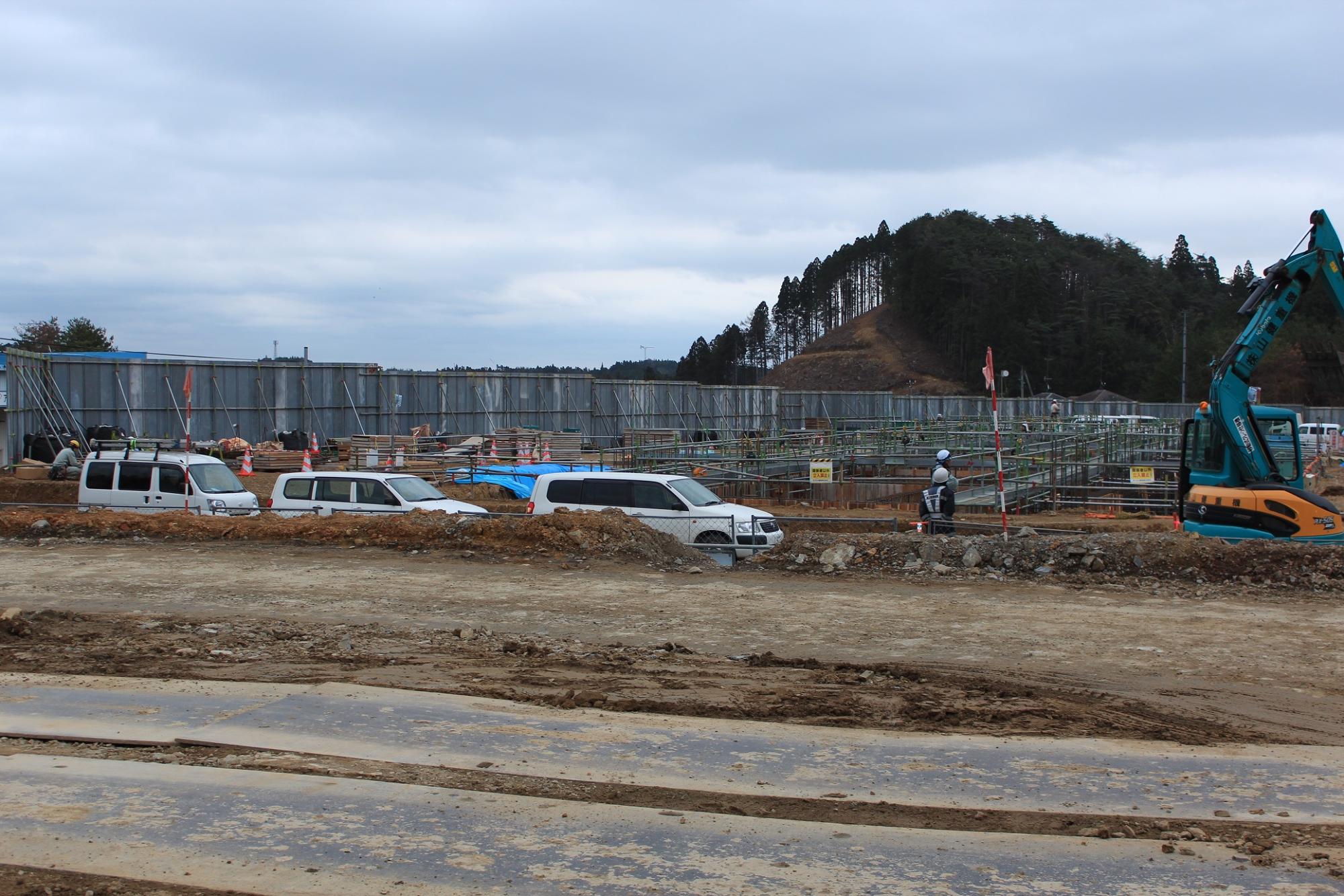 複数の白い車両と作業員が行き交う広い建設現場で、鉄骨の骨組みや足場が組まれた構造物の建設が進み、背景には木々の生い茂った小高い山が見える写真