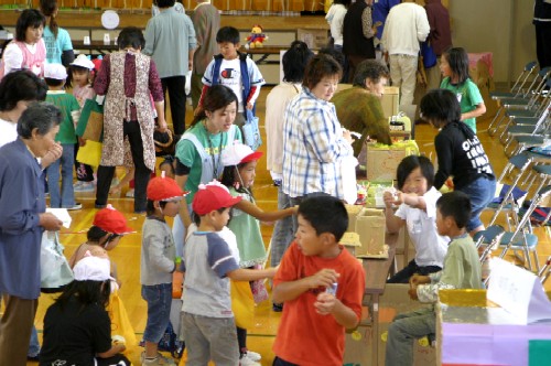 広々とした体育館の中で、長机が並び、コーナーが設けられ、子どもたちが列を作って順番を待ちながら周囲の友達とはしゃぎ、大人たちがその様子を穏やかな表情で見守っているうなばらまつりの写真