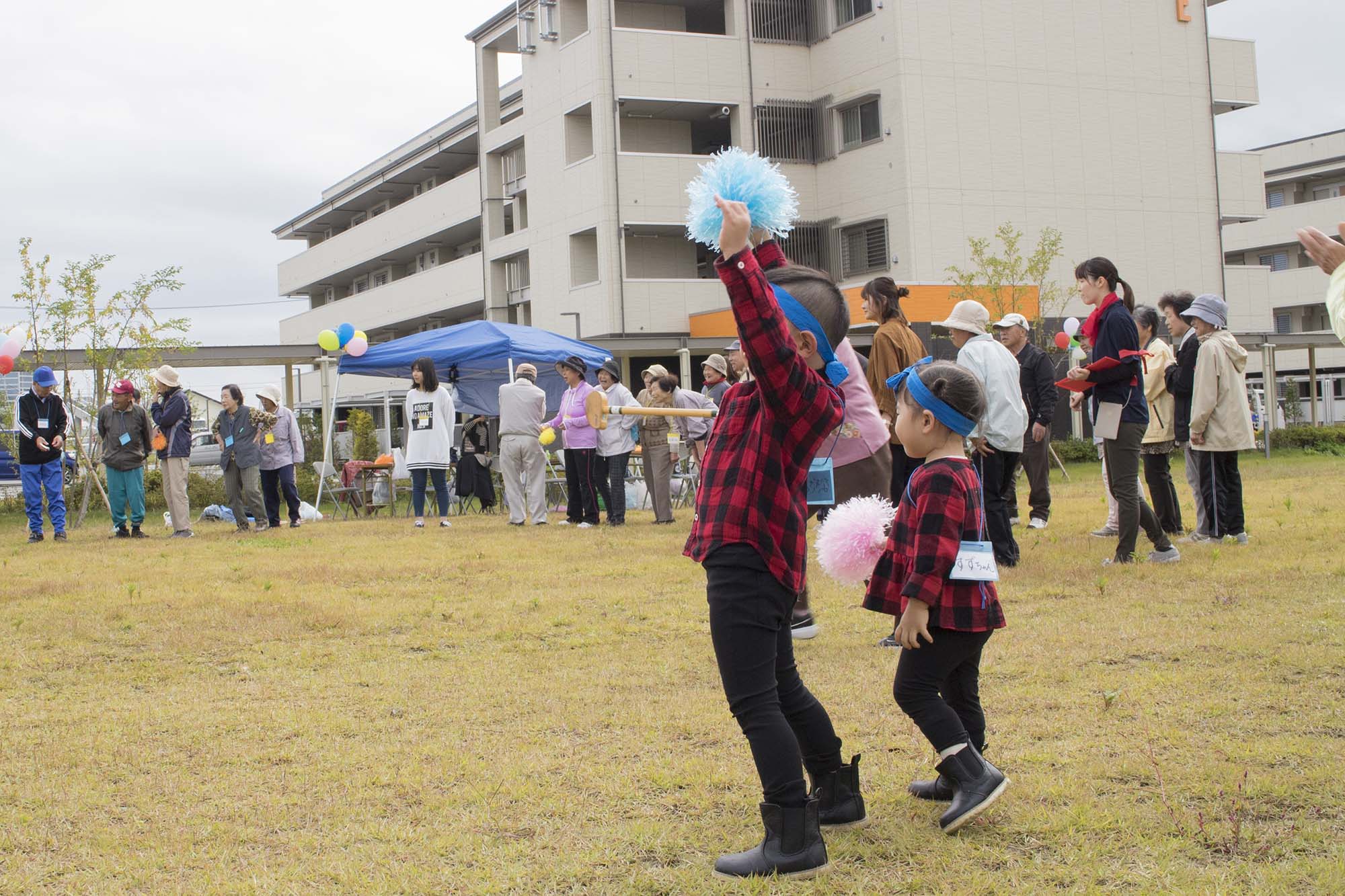 広場にたくさんの人が集まり、赤と黒のチェックの服を着た子どもが青いポンポンを掲げて応援し、背景には高齢者や大人が並んでいる地域の運動会の写真