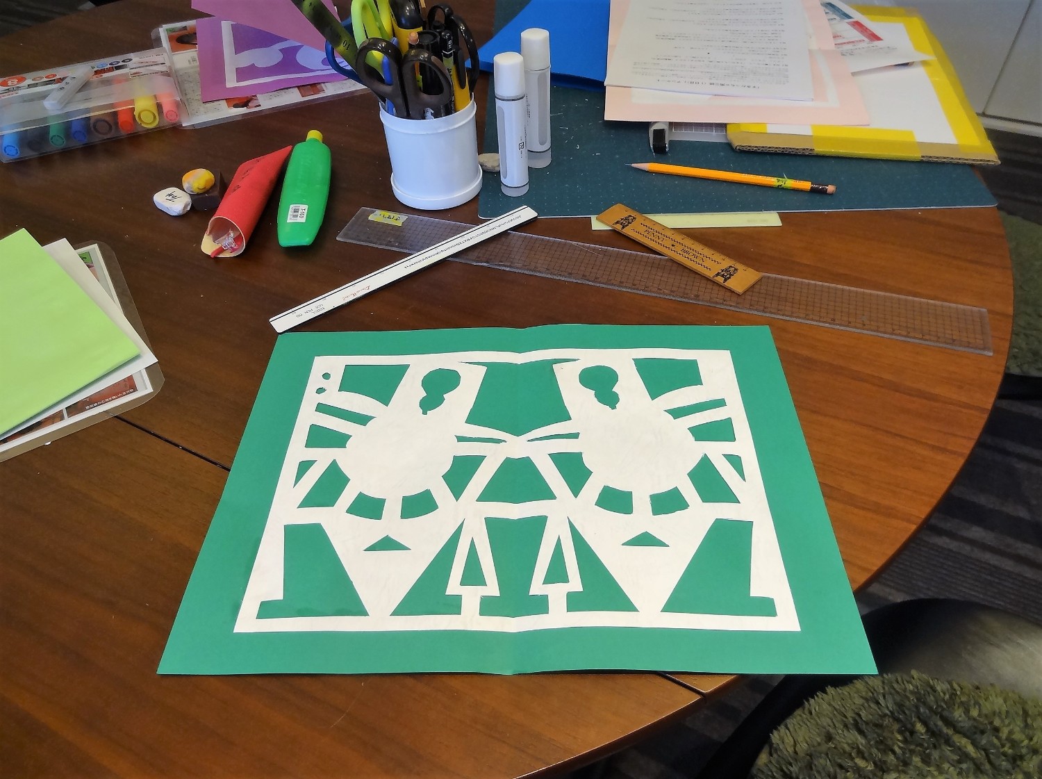 机の上に緑色の台紙に貼られた白い切り絵が置かれ、その周囲にはハサミやのりなどの文房具と、赤・青・黄色などのカラフルな紙が置かれている写真