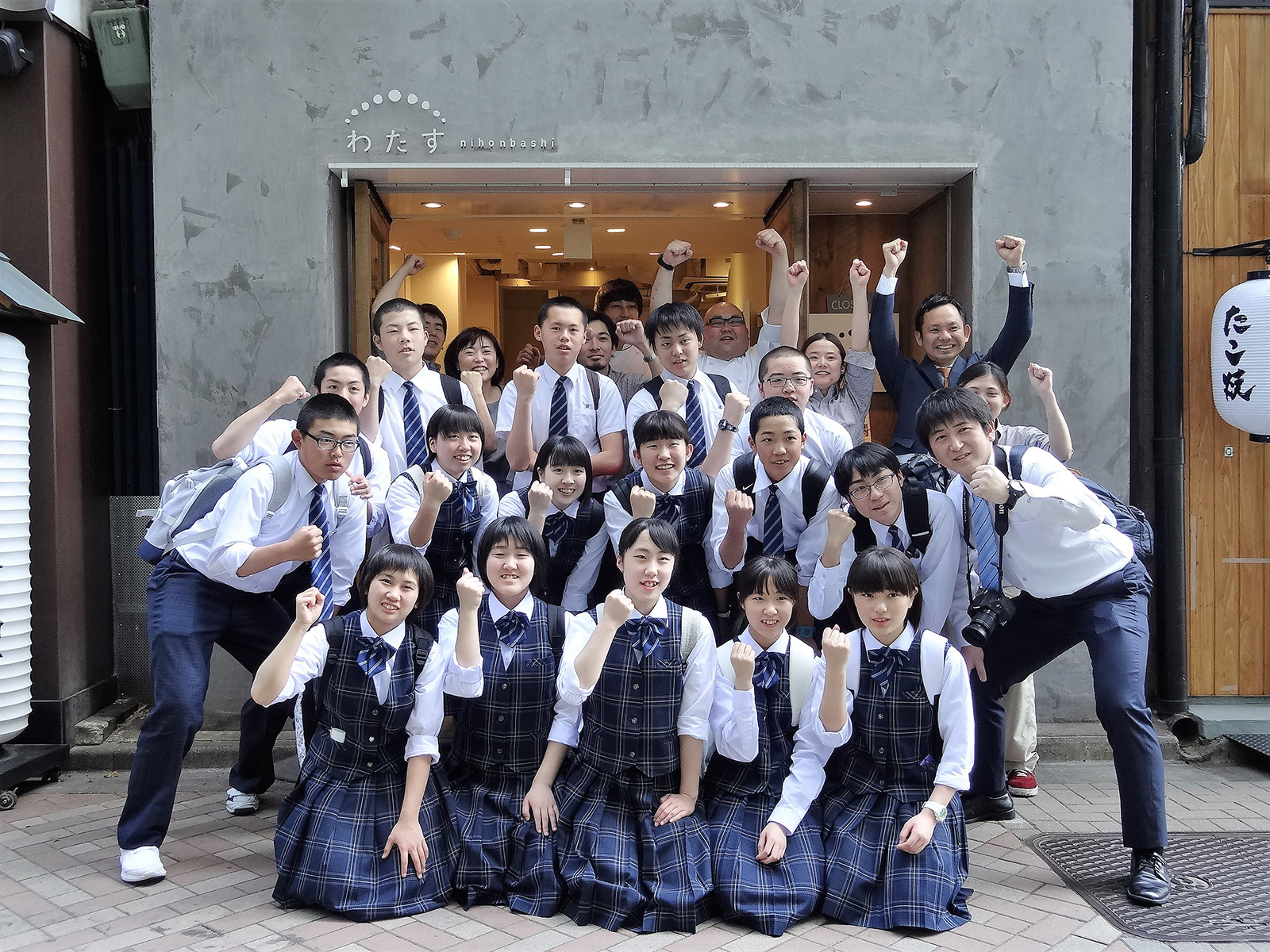 「わたす」の文字が入り口前に掲げられたグレーの建物の前で、生徒と引率の先生など、全員が拳を見せたポーズをとって写した集合写真