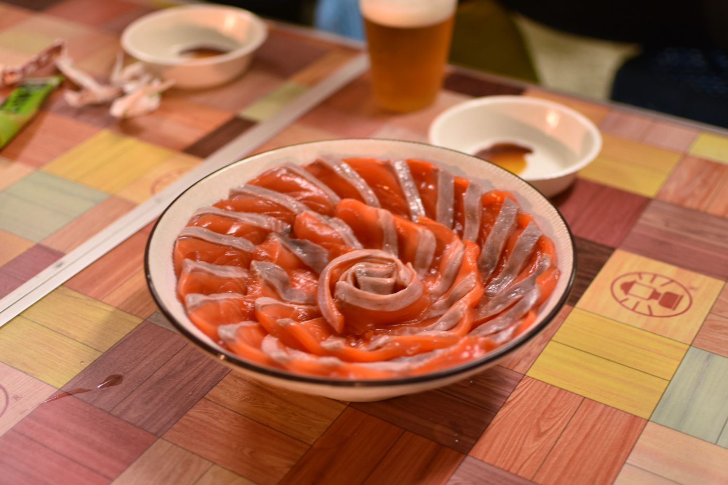 銀鮭の刺身が花のように盛りつけられた皿がテーブル中央に置かれ、その周囲に醤油皿とビールが並んでいる写真
