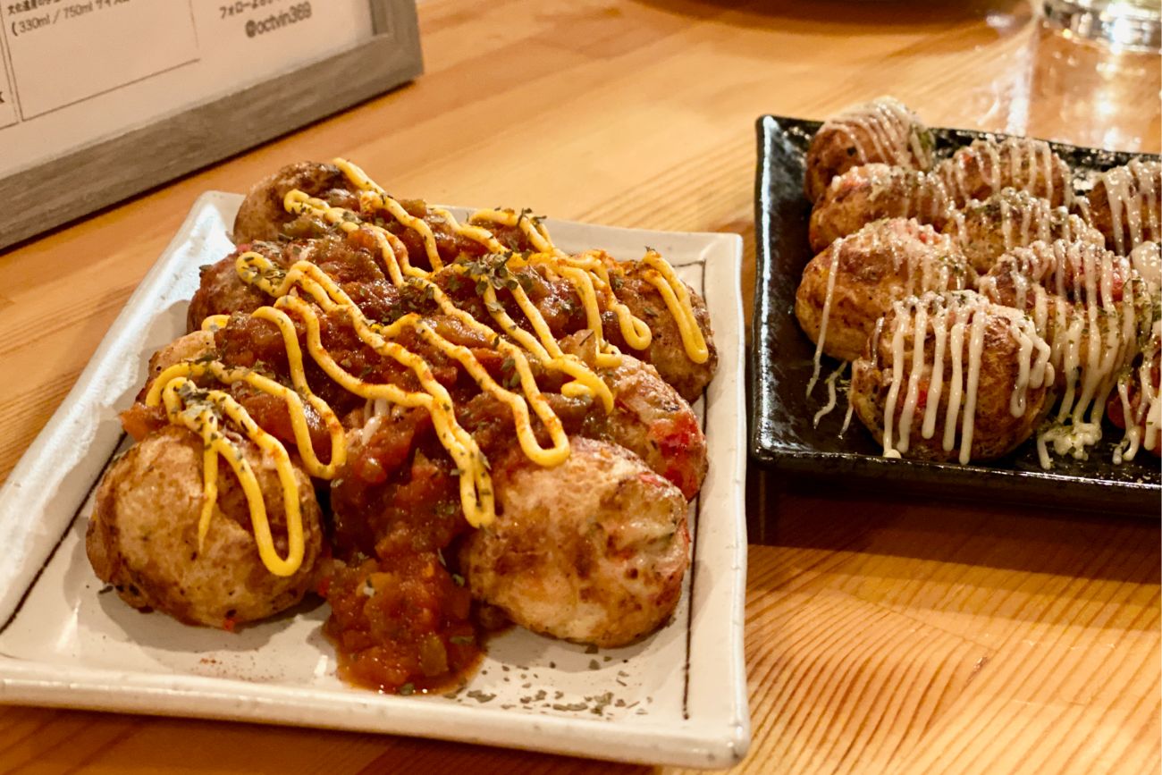 白い皿にチリソースとマヨネーズがかけられ、黒い皿にマヨネーズがかけられた2種類のたこ焼きが盛り付けられている料理の写真