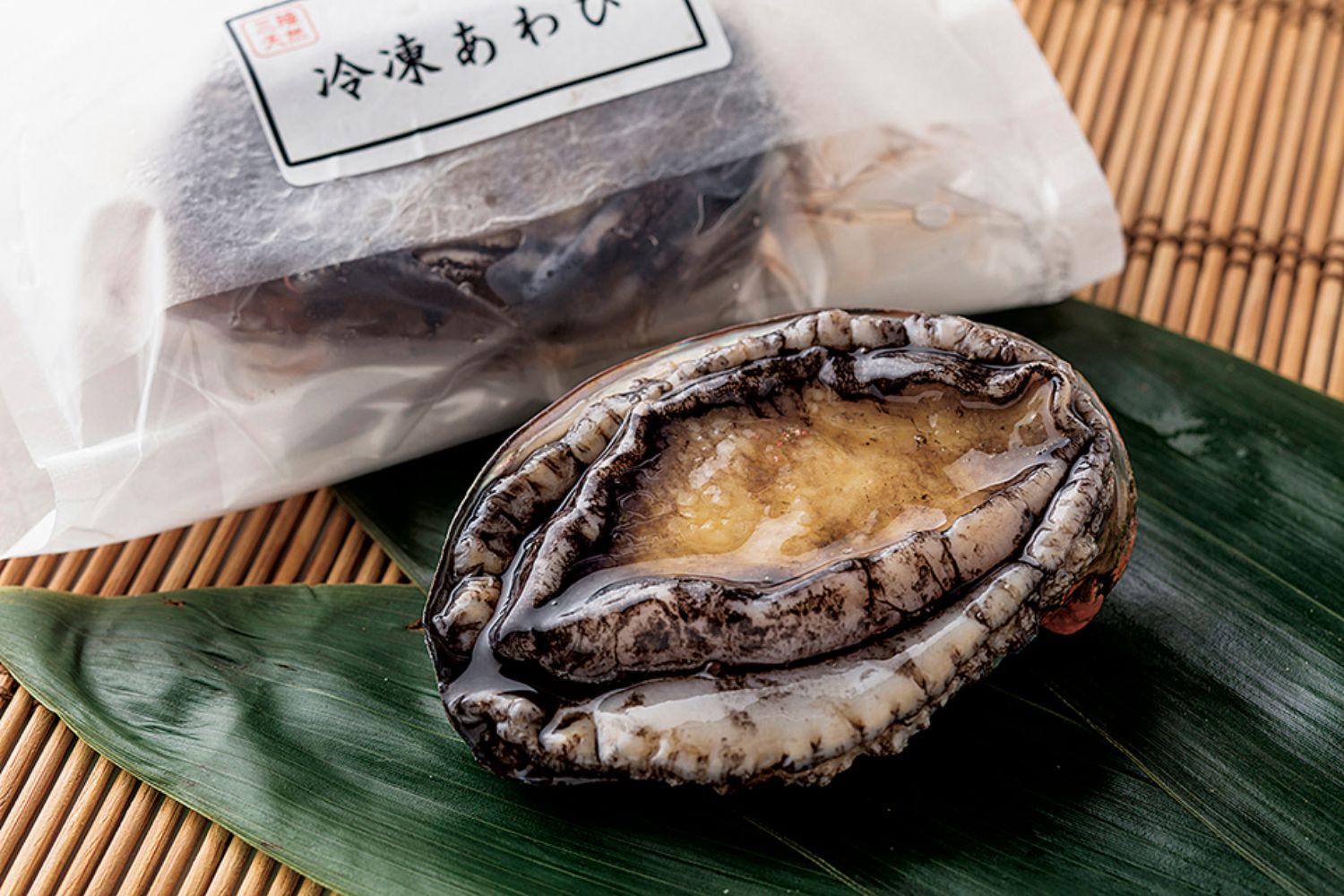 黒と白のまだら模様を持つアワビが、緑色の笹の葉の上に乗せられ、後ろには「冷凍あわび」と書かれたパッケージが写っている写真
