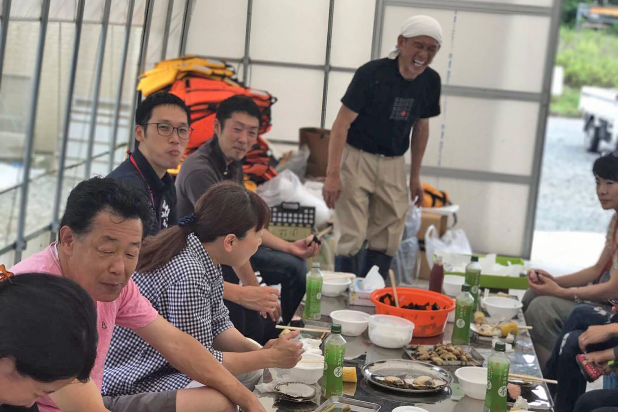 複数人がテーブルを囲んで楽しそうに食事をしながら談笑しており、海産物の串焼きやお茶などが並んでいる写真