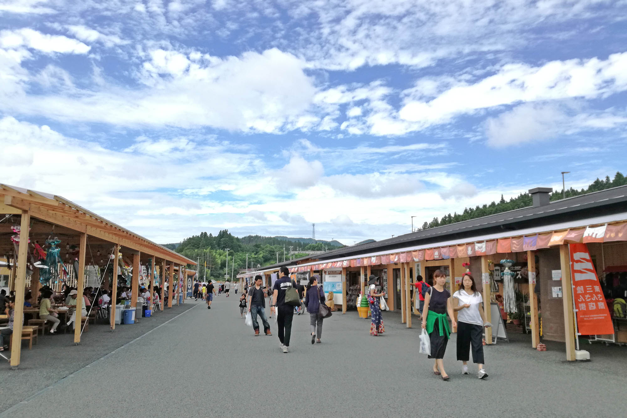 晴れた青空の下で、多くの人々が木造の商店街を散策したり買い物したりしている写真