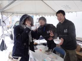 テントの下で男女3人が湯気が上がるお椀を持ってポーズをとっている写真