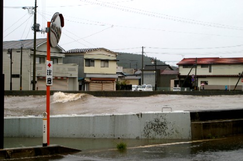 分厚い雲に覆われた天候の中、白いコンクリートの護岸の上まで濁流が迫り、波打っている川の様子と、川沿いには多くの住宅が立ち並び、手前の道路には浸水した水が溜まっている災害時の河川の様子の写真
