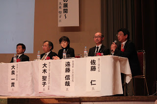 舞台上にそれぞれの前には名札が置かれ舞台上の机に座っているパネリスト5名の皆さんの写真