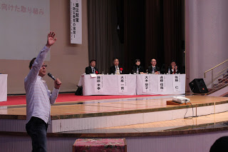 舞台下の客席でマイクを持って話をしている男性と、舞台上の机に座っているパネリスト5名の方々が写っている国土政策フォーラムの様子を写した写真