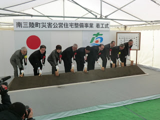 「第三期市営住宅建設事業着工式」の横断幕と国旗、町旗が掲げられ、テント下で数人の関係者が並んで鍬入れをしている様子の写真