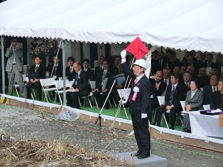 多くの関係者の方々がテント下の来賓席に座って見守る中、立木伐採の合図の為、スーツ姿の男性が赤い旗を頭上に挙げて立っている様子の写真