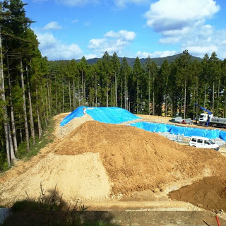 青いシートで覆われた土の山が見え、周囲にトラックや重機が見えている写真