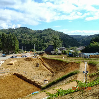 土を削って作られた傾斜のある土地に白い排水管が通されており、まわりには集落や山が見える工事現場の写真
