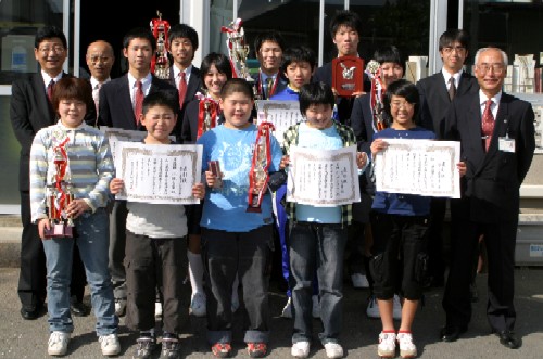 子どもたちが大会で獲得した賞状とトロフィー、楯を手に中央に並んで立ち、両隣に立っているスーツ姿の関係者たちとともに皆が笑顔で記念撮影に応じている集合写真