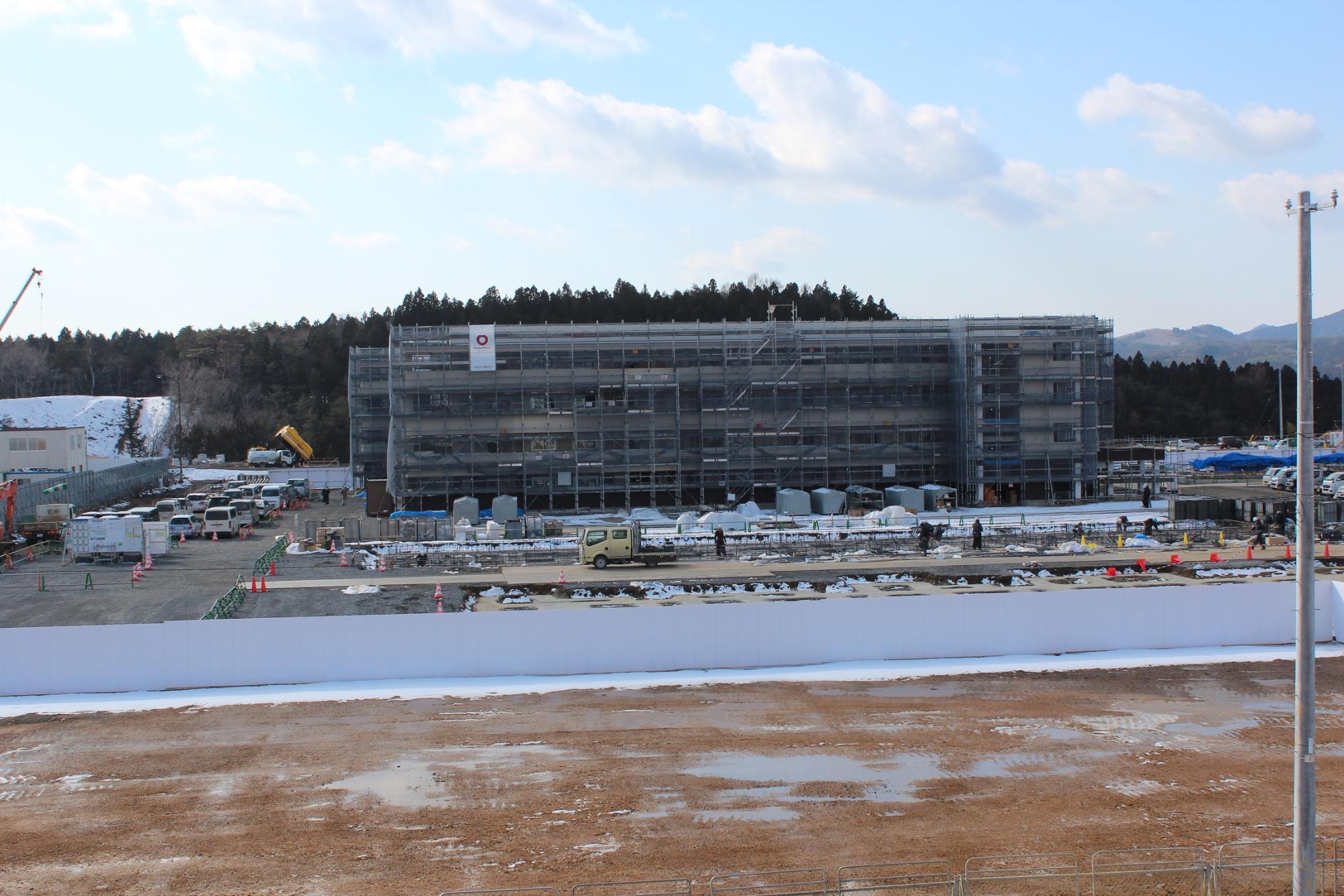 雪が部分的に残る地面の奥に、足場に囲まれた中層の建設中の建物があり、その周囲で作業員や車両が活動している冬の工事現場の写真