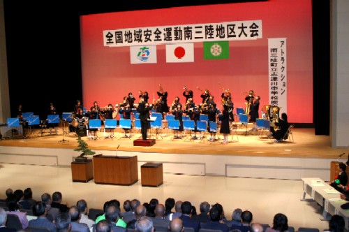 「全国地域安全運動南三陸地区大会」の大きな横断幕が掲げられたホールステージで、制服姿の生徒たちが多数の観客を前に、指揮者に合わせて楽器を演奏している様子の写真